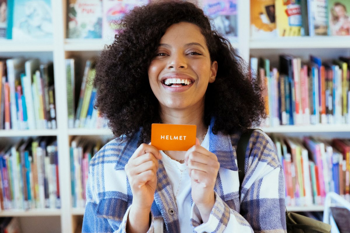 A smiling person stands in front of a book shelf holding an orange library card.