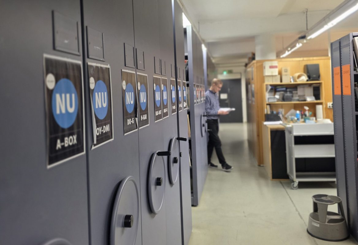 Etualalla musiikkivaraston arkistohyllyt. Takana häämöttää työtiloja ja työntekijä.